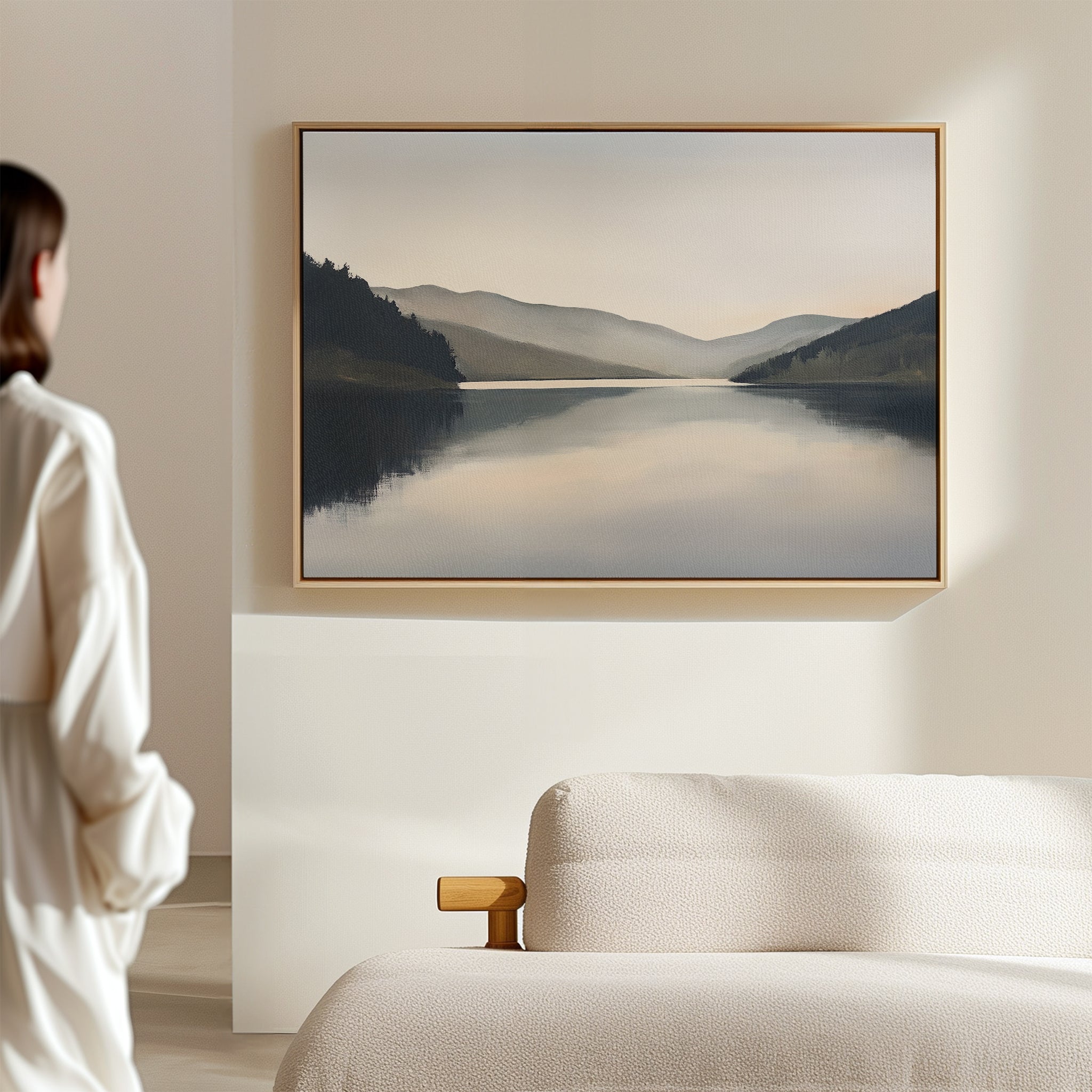 A woman in a white robe stands in a minimalist bedroom, gazing at Mirror Valley Canvas Art - Horizontal, a framed landscape above the bed that enhances the space with its serene natural palette.