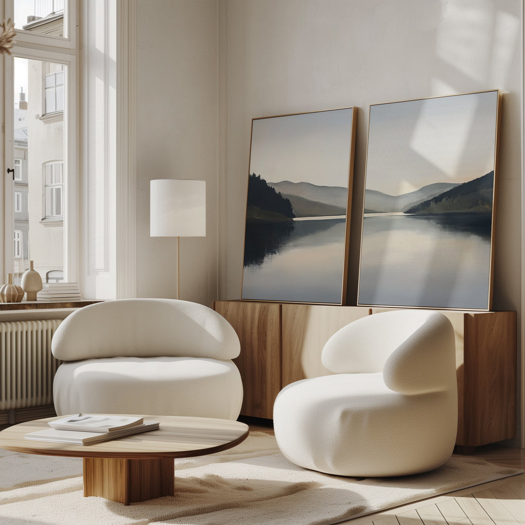 A modern living room features two rounded white armchairs, a low wooden coffee table, and Mirror Valley - Set of 2 landscape canvas prints on a wood sideboard, with natural light streaming in through tall windows.