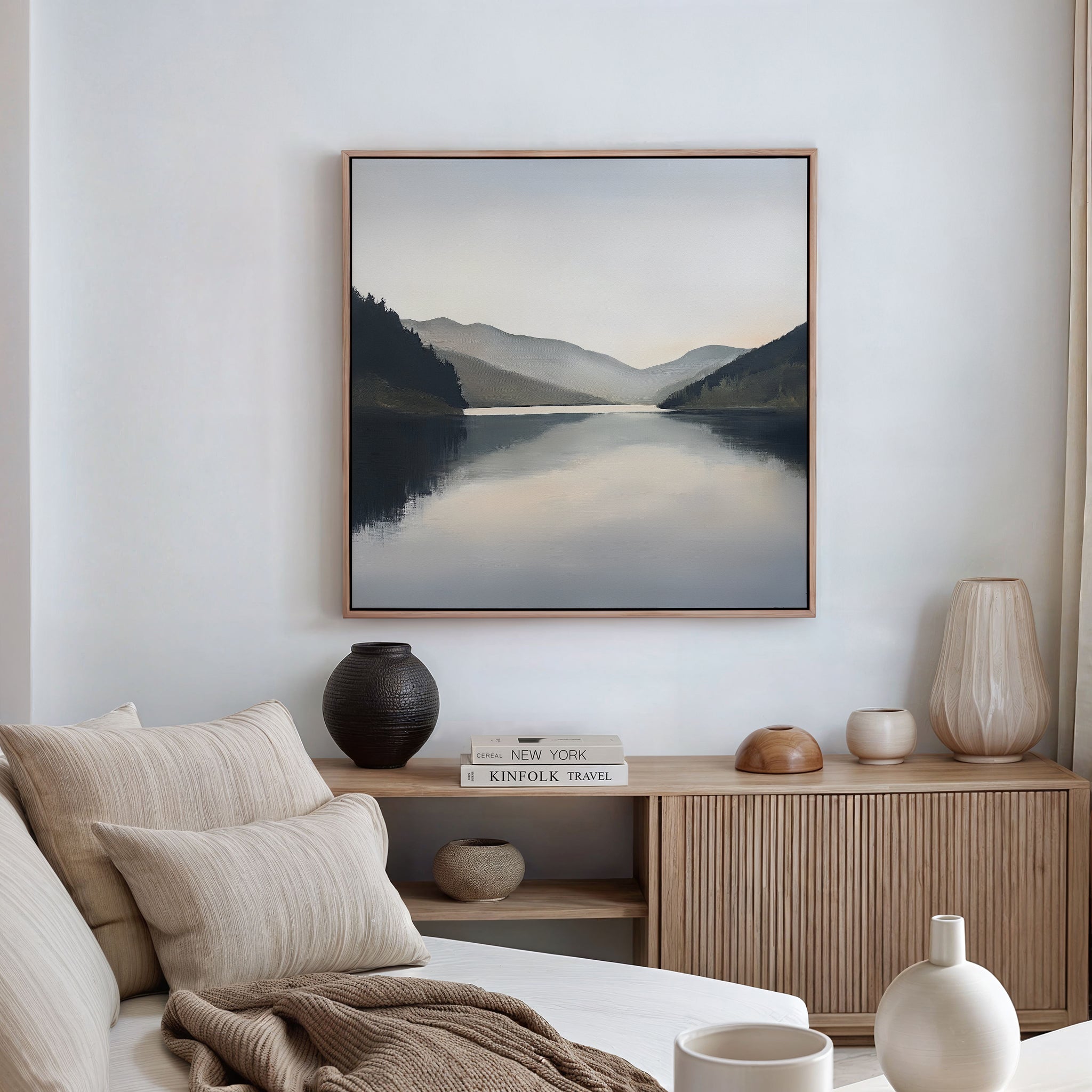 A modern living room with a beige sofa, neutral-toned cushions, wooden cabinet, decorative vases, and the Mirror Valley Canvas Art - Square featuring a tranquil lake and mountains as the centerpiece wall décor.