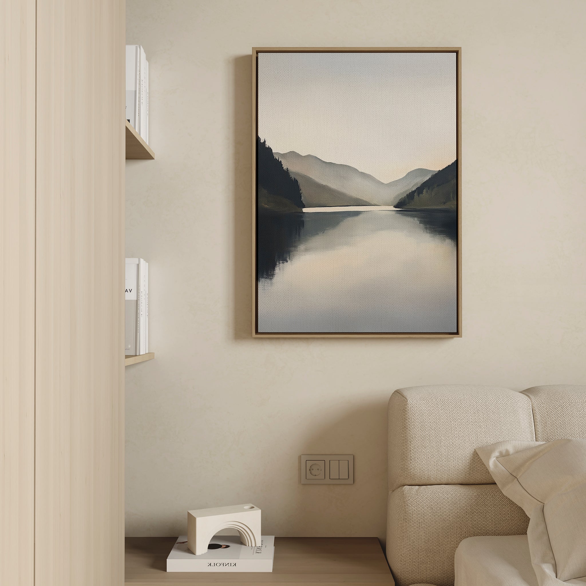 The Mirror Valley Canvas Art - Vertical, featuring a serene lake and mountain landscape, is framed and hangs on a beige wall above a light sofa. To the left, there’s a bookshelf and Nordic decor visible in the room.