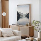 A serene living room features a light wood sideboard, Nordic accents, neutral vases, stacked books, a green plant, white sofa, round pendant light, and the Mirror Valley Canvas Art - Vertical depicting mountains and a lake.