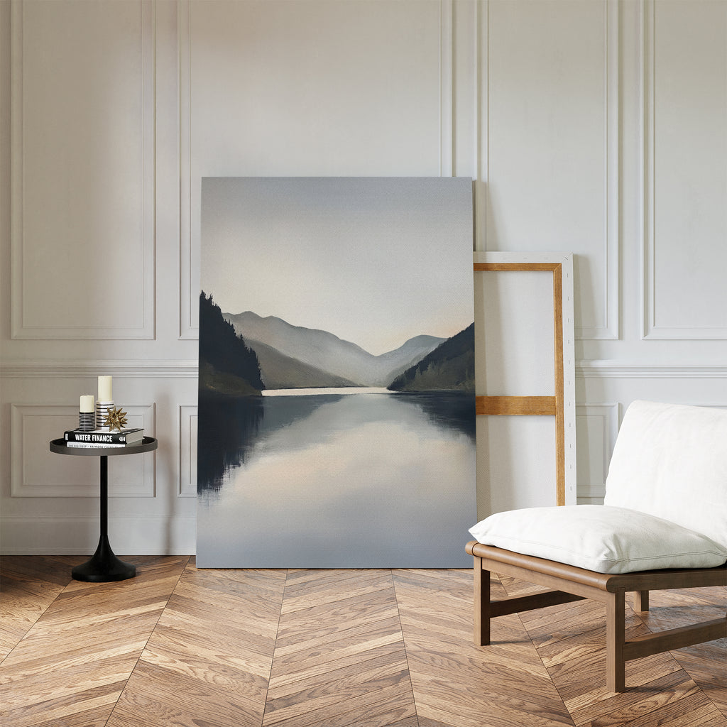 A minimalist room with wood herringbone floors, white walls, Nordic decor, a cushioned chair, a small round table with candles and books, and the Mirror Valley Canvas Art - Vertical of mountains and a lake resting against the wall.