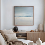 A serene living room features the Misty Morning Abstract Canvas Art in blue and white above a wooden sideboard, with neutral vases, books, and a beige sofa with striped cushions, creating a tranquil atmosphere.
