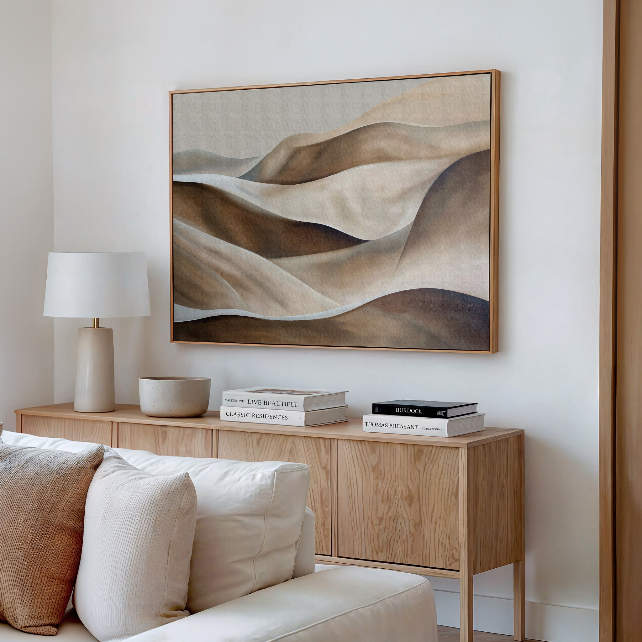 A minimalist living room features a wooden sideboard, neutral decor, stacked books, a lamp, and the Desert Waves Abstract Canvas Art with soft earthy tones, hanging above a cream-colored sofa on a white wall.