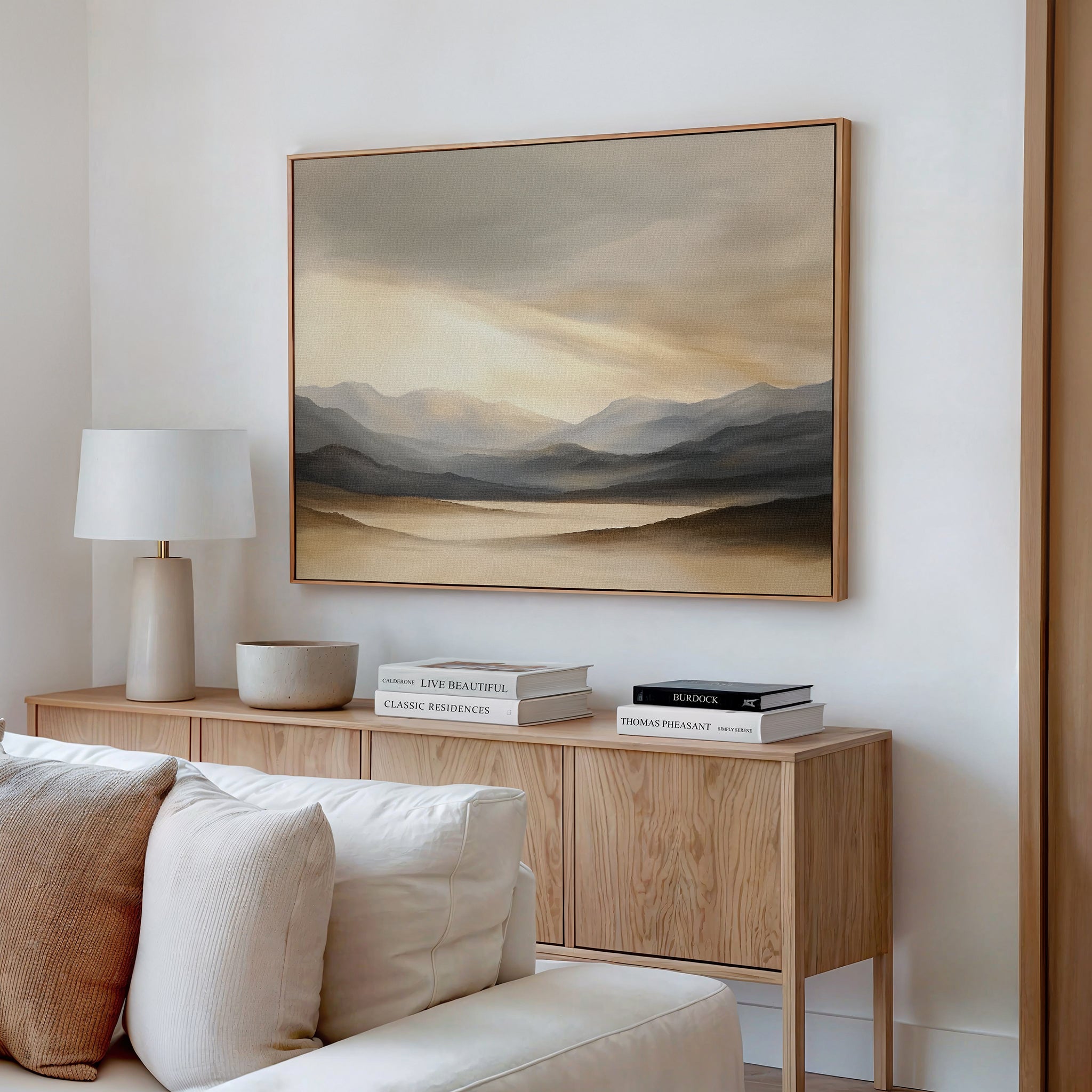 A modern living room with a light wood sideboard, white lamp, bowl, stacked books, and the Mountain Reverie Canvas Art - Horizontal—a neutral-toned landscape—hanging above a cream-colored sofa.