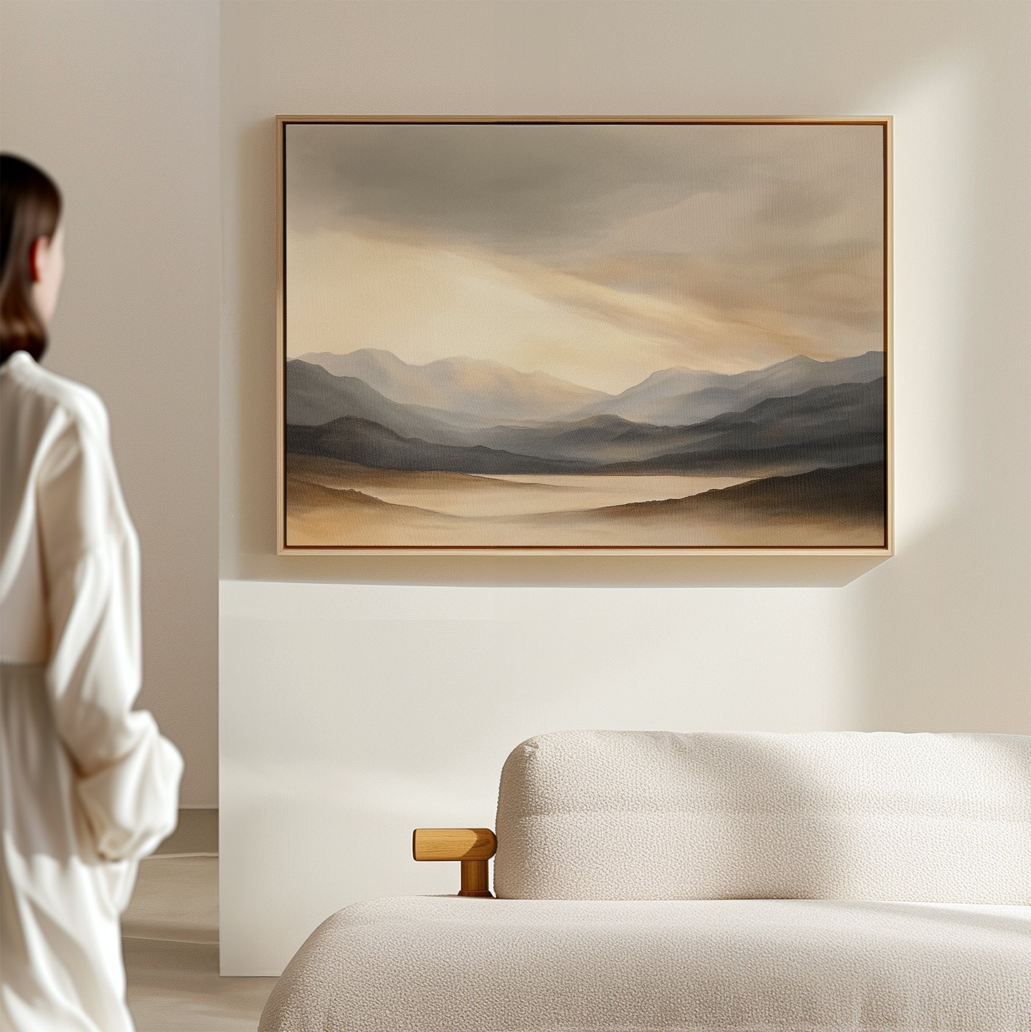 A person stands in a softly lit room, gazing at the Mountain Reverie Canvas Art - Horizontal above a beige sofa. The neutral tones and minimalist decor evoke the calm, natural beauty of a tranquil Nordic Park.