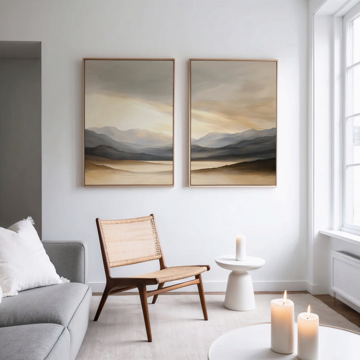 A modern, minimal living room features a light gray sofa, wooden chair, small round white table with candles, and two "Mountain Reverie - Set of 2" landscape paintings on a white wall. Sunlight streams through a large window.
