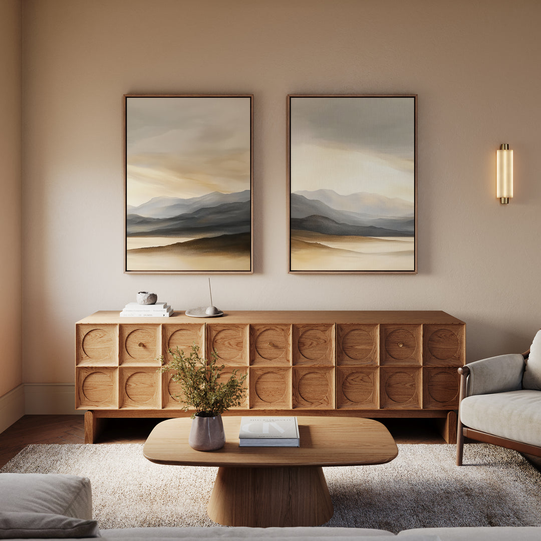 A modern living room featuring a wooden coffee table, textured sideboard, plush armchair, wall sconce, a vase with greenery on the table, and the Mountain Reverie - Set of 2 serene art prints displayed above.