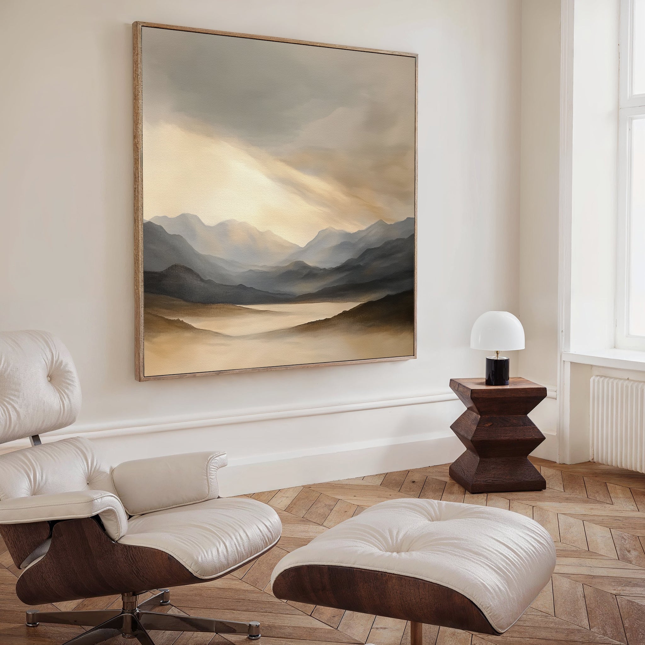 A modern living room with a cream lounge chair and ottoman, a dark wood side table topped with a white lamp, and the "Mountain Reverie Canvas Art - Square," featuring misty mountains and a river, displayed on the beige wall.