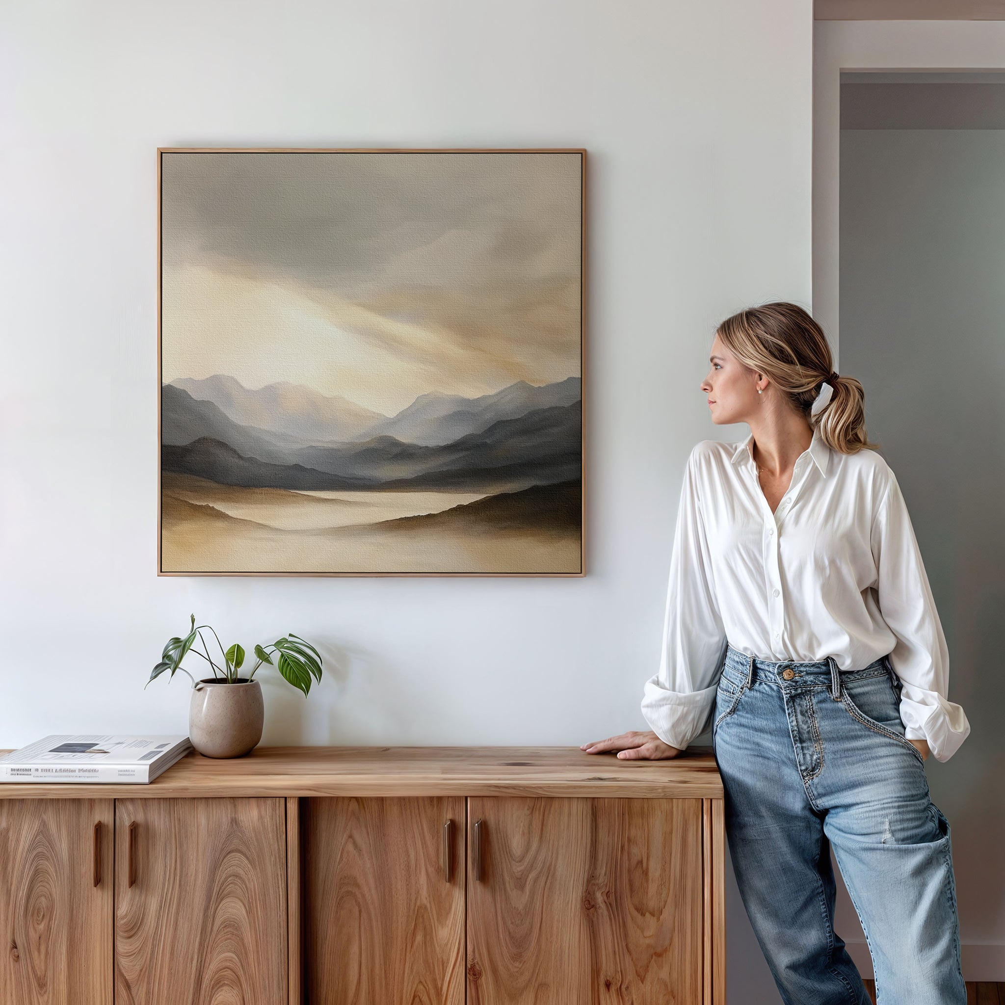 A woman in a white shirt and jeans leans against a wooden cabinet, admiring the Mountain Reverie Canvas Art - Square. A potted plant and book on the cabinet add to the calm, minimalist decor of the room.
