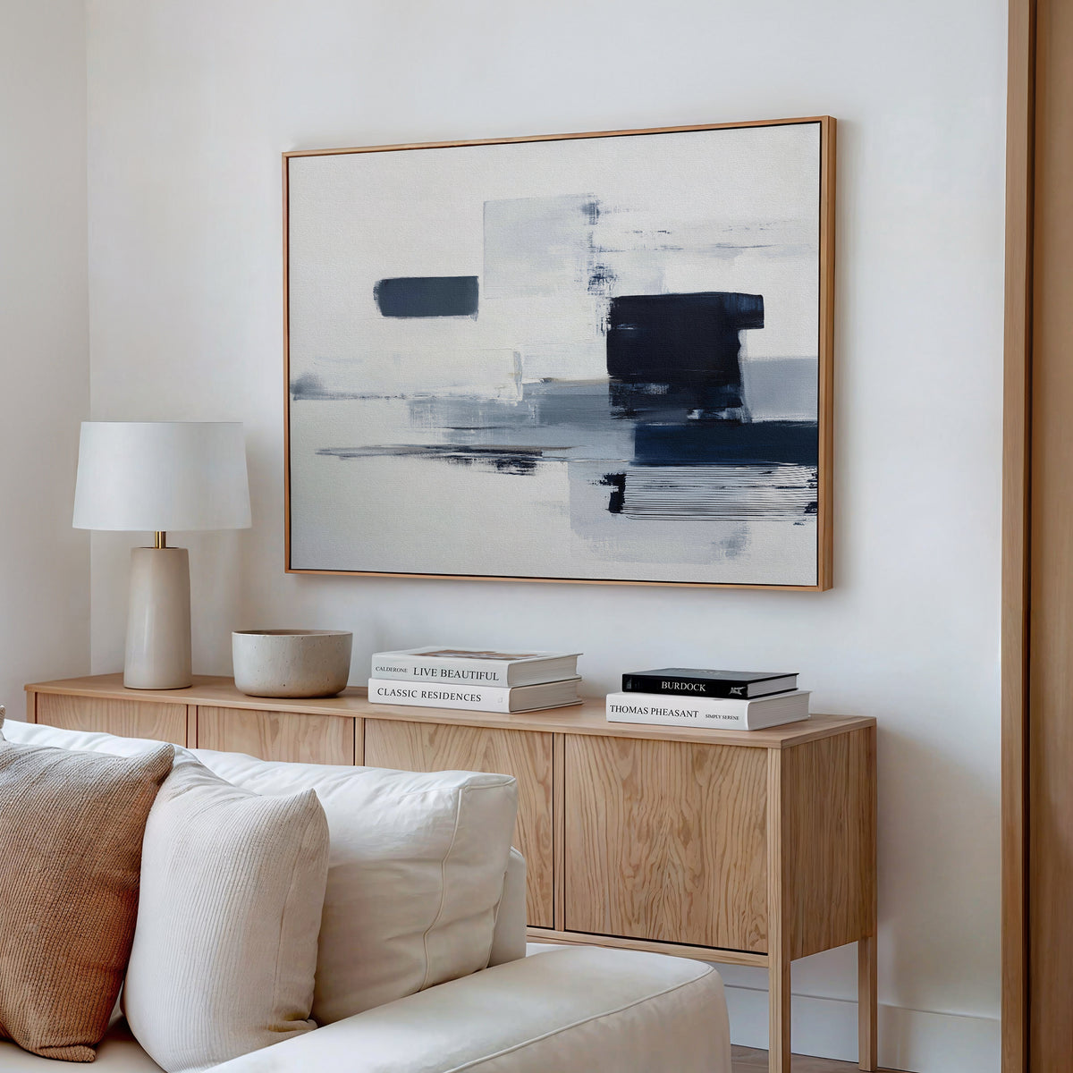 A modern living room features a light wood sideboard, white table lamp, stacked books, and a bowl. Above the beige sofa on a white wall hangs the Navy Brushwork Canvas Art - Horizontal, adding depth and intrigue to the decor.