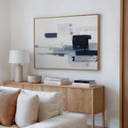 A modern living room features a light wood sideboard, white table lamp, stacked books, and a bowl. Above the beige sofa on a white wall hangs the Navy Brushwork Canvas Art - Horizontal, adding depth and intrigue to the decor.