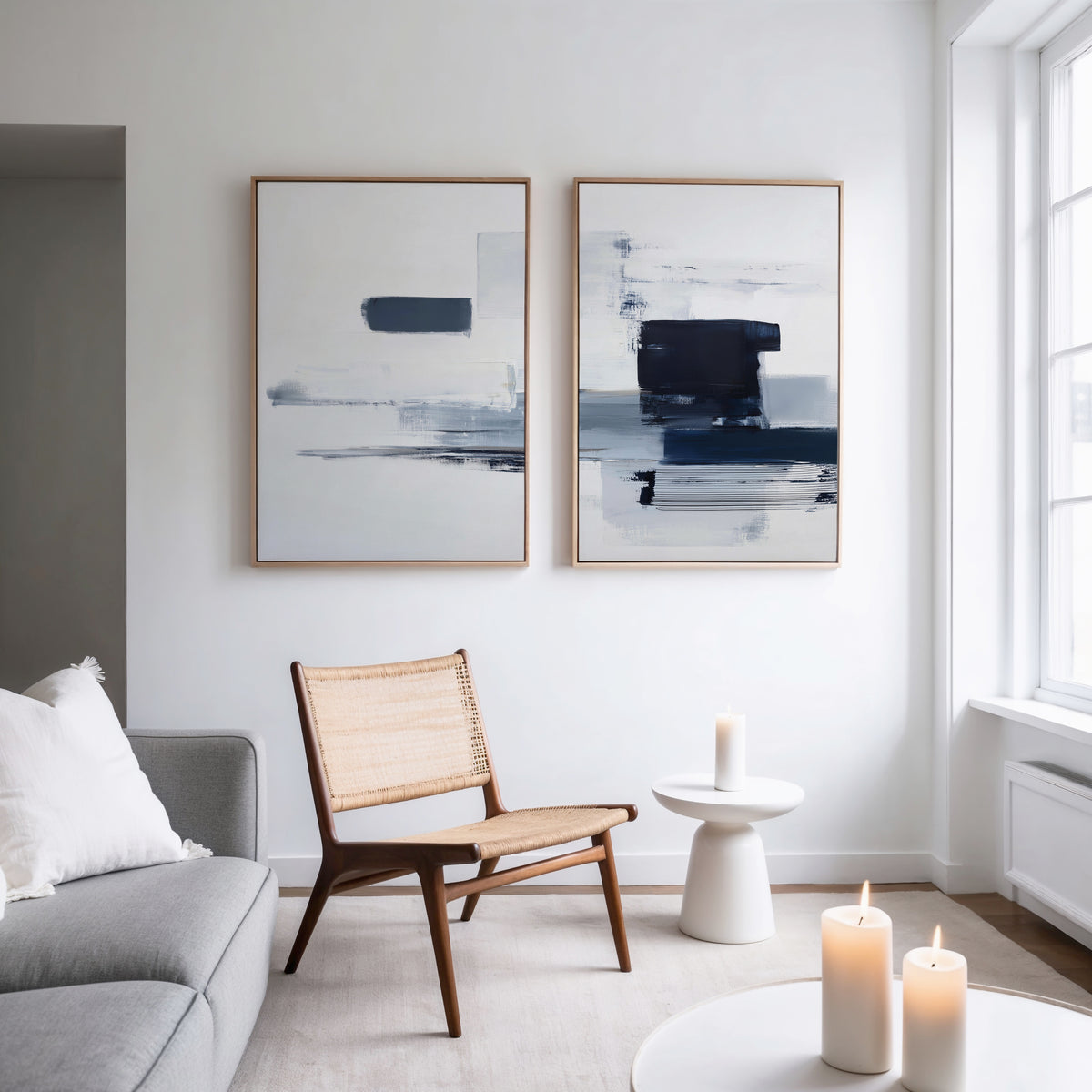 A modern, minimal living room features a woven chair, round side table, lit candles, light gray sofa, and the Navy Brushwork - Set Of 2 wall art on a white wall by a large window—ideal for minimalist home decor fans.