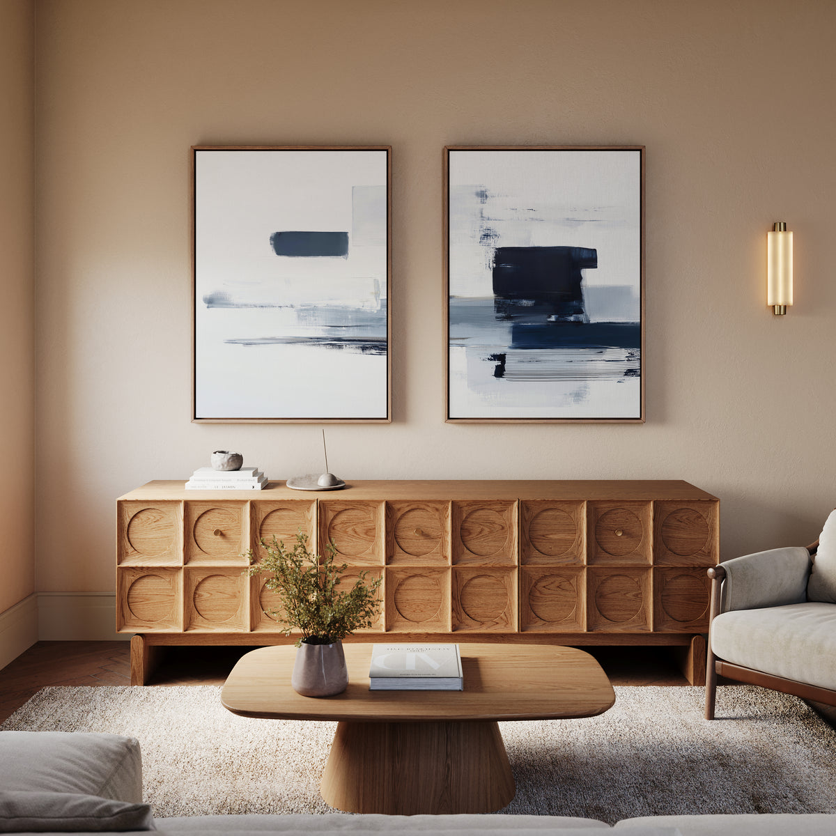 A modern living room featuring a wooden sideboard, Navy Brushwork - Set Of 2 wall art, a light wood coffee table with a vase of greenery and a book, and a beige armchair illuminated by soft, warm lighting.