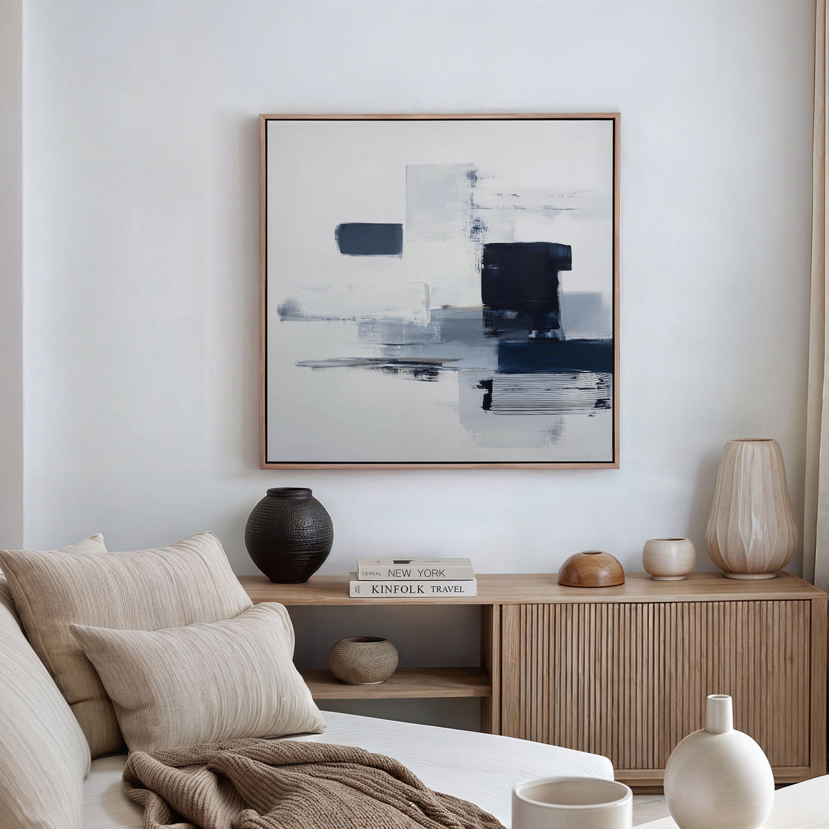 A modern living room features a beige sofa, neutral cushions, a wooden sideboard with vases and books, and the Navy Brushwork Canvas Art - Square on a white wall for a minimalist home decor touch.