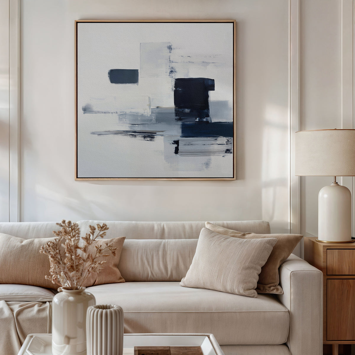 A modern living room features a beige sofa, cream cushions, a side table with a lamp, and dried flowers in a vase. Above the sofa, the Navy Brushwork Canvas Art - Square brings flair to the minimalist home decor aesthetic.