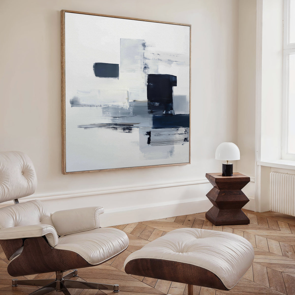 A modern living room with a white leather lounge chair and ottoman, a small wooden side table with a white lamp, and the Navy Brushwork Canvas Art - Square adding sophisticated style to this minimalist home decor.