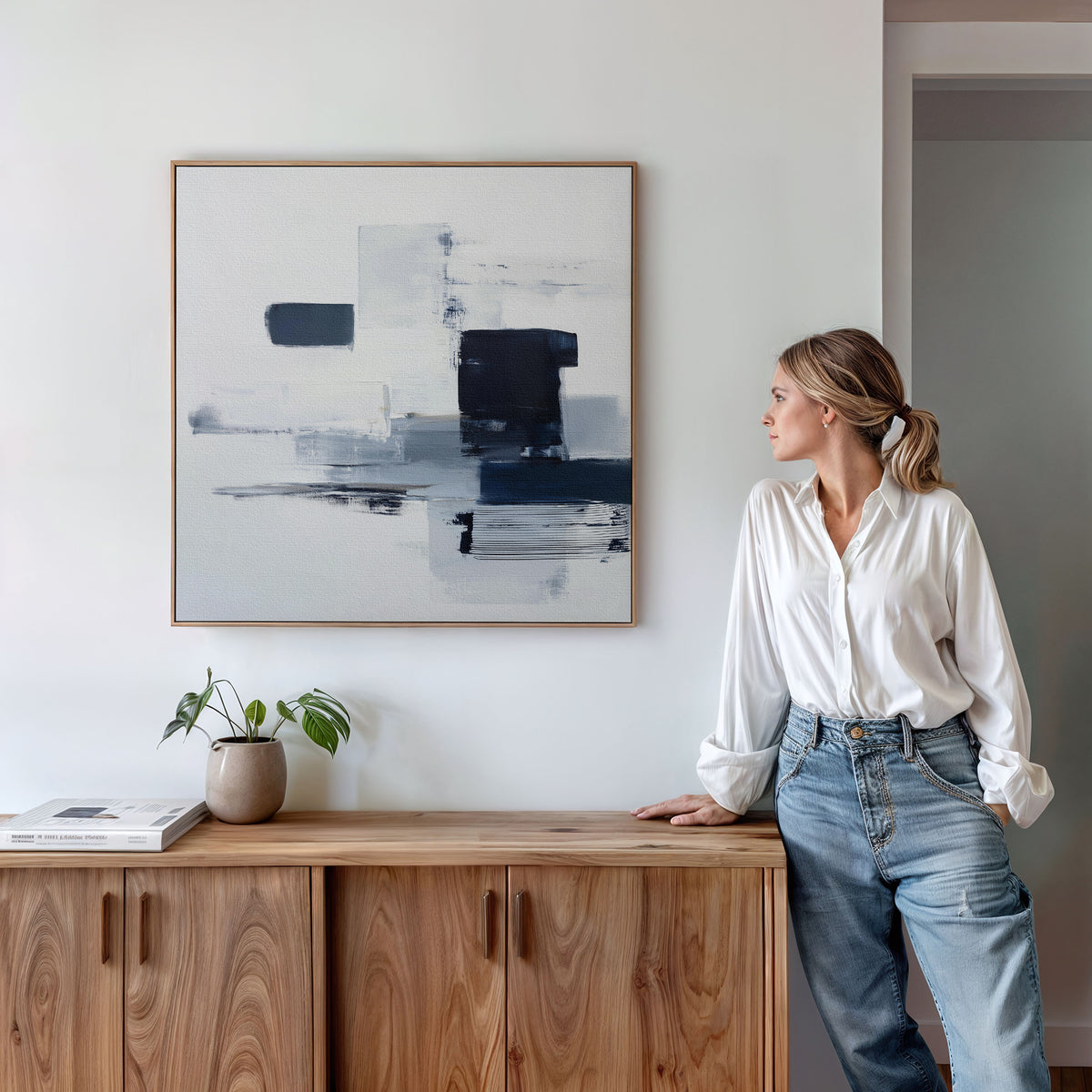A woman in a white blouse and jeans leans against a wooden cabinet, admiring the Navy Brushwork Canvas Art – Square on the wall. Minimalist decor includes a small plant and books atop the cabinet.