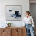 A woman in a white blouse and jeans leans against a wooden cabinet, admiring the Navy Brushwork Canvas Art – Square on the wall. Minimalist decor includes a small plant and books atop the cabinet.