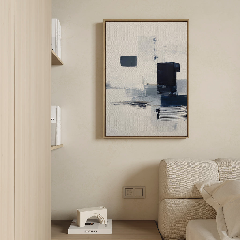 A modern living room features a light sofa, wooden shelves with white books, and the Navy Brushwork Canvas Art - Vertical in blue, white, and black on a beige wall—perfect for minimalist decor.