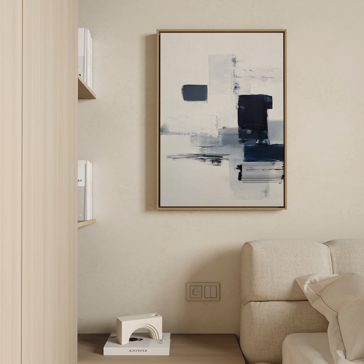 A modern living room features a light sofa, wooden shelves with white books, and the Navy Brushwork Canvas Art - Vertical in blue, white, and black on a beige wall—perfect for minimalist decor.