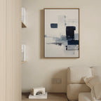 A modern living room features a light sofa, wooden shelves with white books, and the Navy Brushwork Canvas Art - Vertical in blue, white, and black on a beige wall—perfect for minimalist decor.
