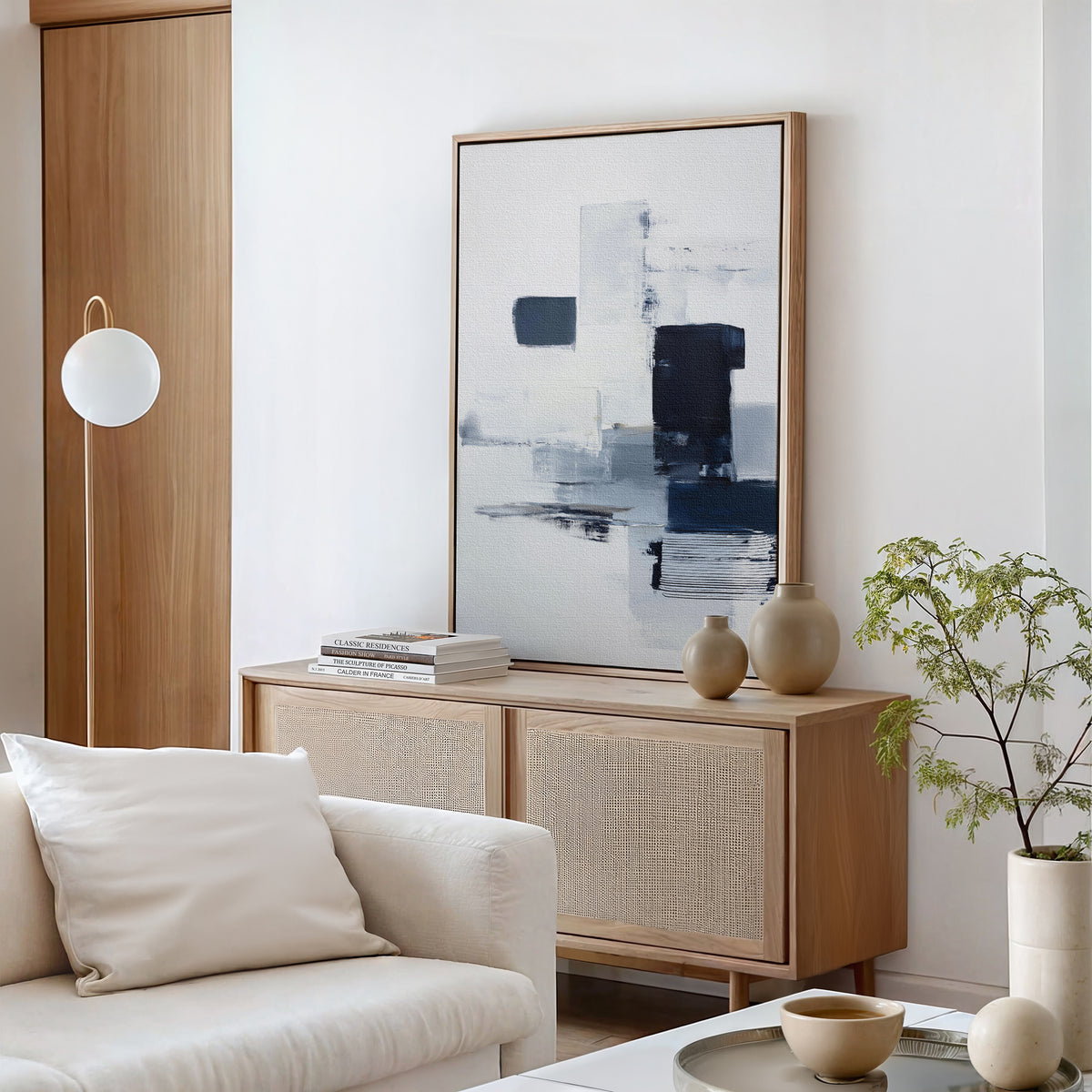 Minimalist living room featuring a light wood sideboard, Navy Brushwork Canvas Art - Vertical, neutral vases, a green plant, and a white sofa with a cream cushion in a bright, modern space—perfect for minimalist decor fans.