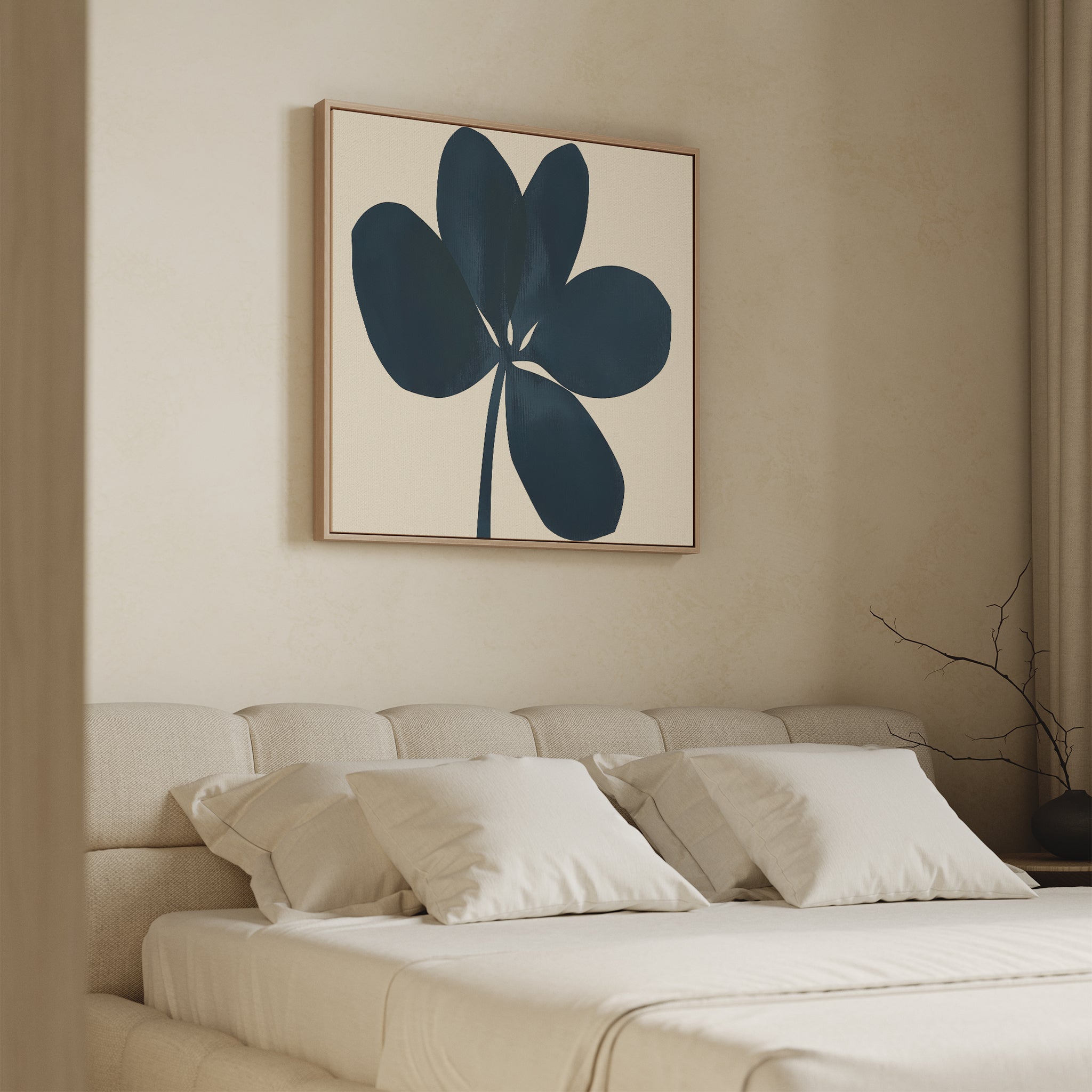 A beige bedroom features a large bed with white bedding and three pillows. Above the bed hangs the Navy Clover Canvas Art - Square, showcasing a dark blue abstract five-petal flower. A bare branch sits on the nightstand.