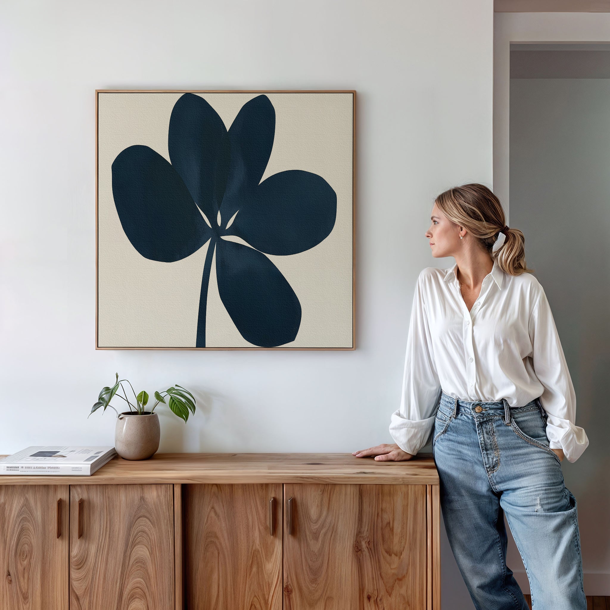 A woman in a white shirt and jeans leans against a wooden cabinet, admiring the Navy Clover Canvas Art - Square, a dark abstract floral piece. Minimalist decor—including a potted plant and books—surrounds her.