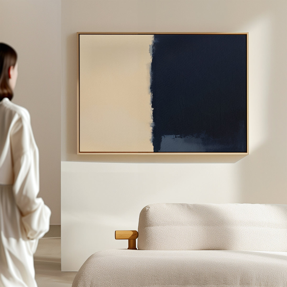 A person in a white shirt stands in a modern, minimal room, admiring the Navy Division Canvas Art - Horizontal—an abstract piece with one half beige and the other dark blue—hanging above a light-colored sofa.