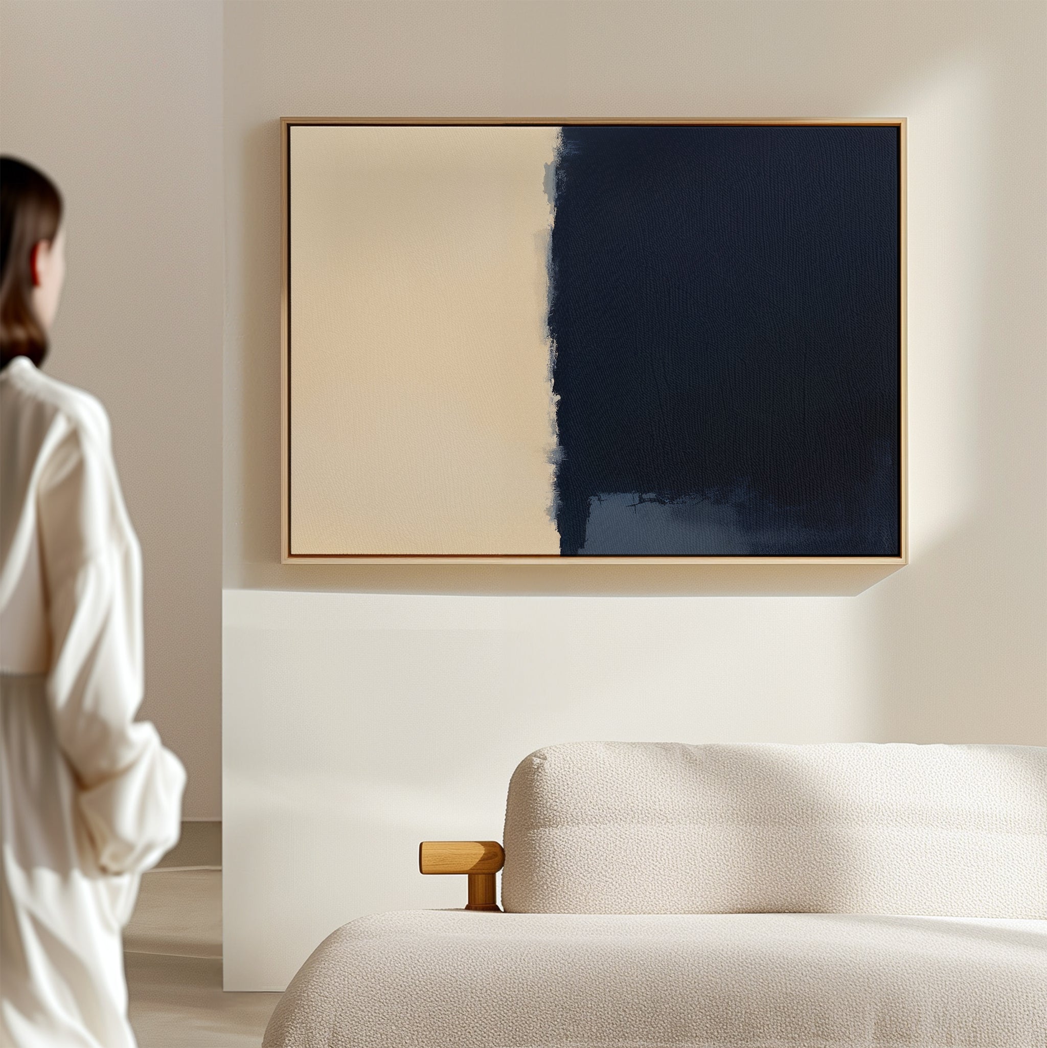 A person in a white shirt stands in a modern, minimal room, admiring the Navy Division Canvas Art - Horizontal—an abstract piece with one half beige and the other dark blue—hanging above a light-colored sofa.