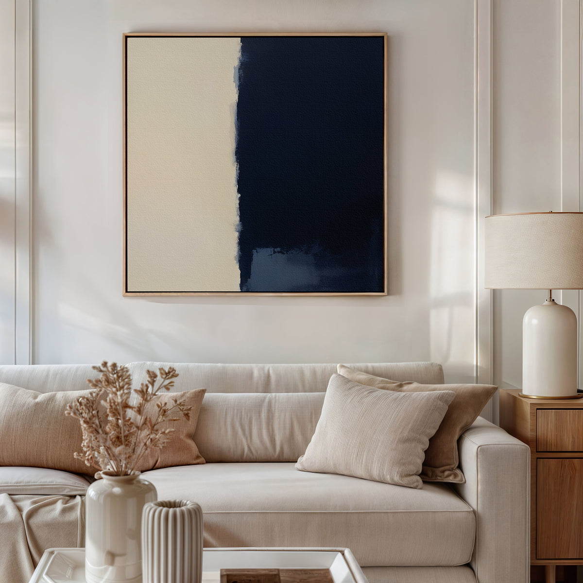 A modern living room features a beige sofa with neutral cushions, a wooden side table with a white lamp, and the Navy Division Canvas Art - Square above the sofa. Dried flowers add charm to the coffee table.
