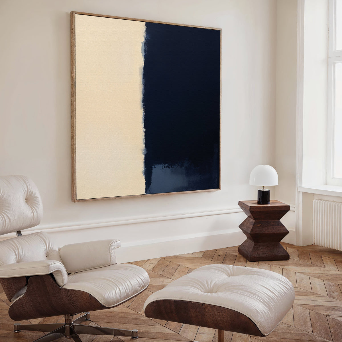 A modern living room featuring a white leather lounge chair and ottoman, a small wooden side table with a dome lamp, and the Navy Division Canvas Art - Square as bold navy blue abstract wall art above.