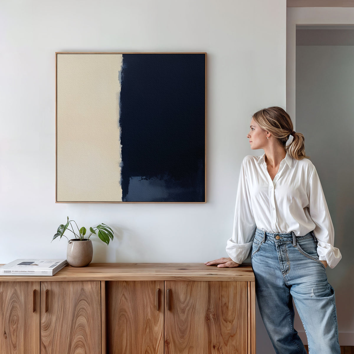 A woman in a white blouse and jeans leans against a wooden sideboard, gazing at the Navy Division Canvas Art - Square, a minimalist navy and cream print framed on a white wall, with a potted plant nearby.