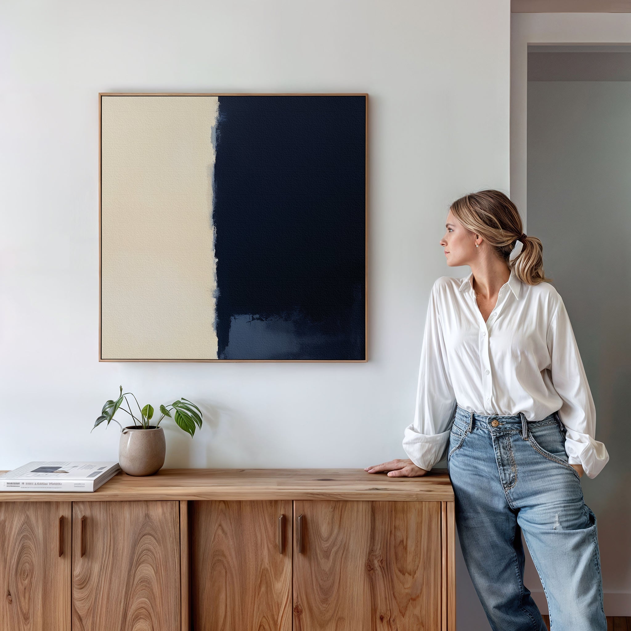 A woman in a white blouse and jeans leans against a wooden sideboard, gazing at the Navy Division Canvas Art - Square, a minimalist navy and cream print framed on a white wall, with a potted plant nearby.