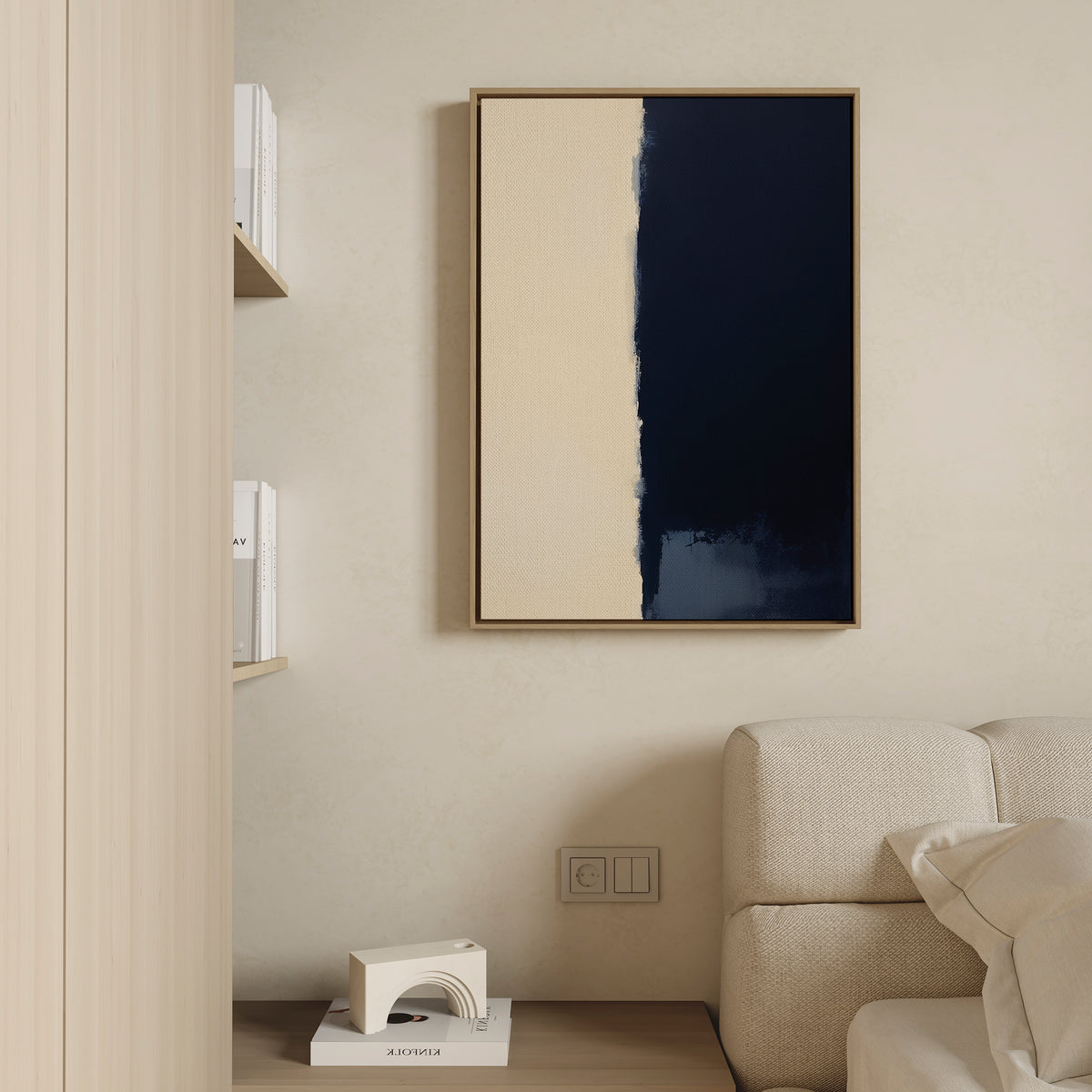 A minimalist living room features a beige wall adorned with the Navy Division Canvas Art - Vertical, showcasing beige and dark blue sections. Nearby, a light-toned couch and a shelf with books and geometric decor exude modern elegance.