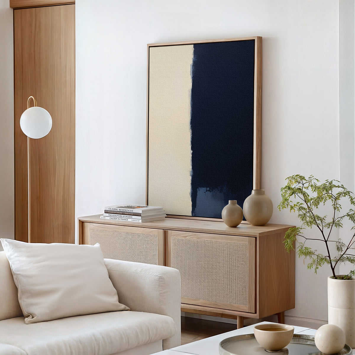 A minimalist living room with a light wood sideboard, neutral decor, a round white lamp, the Navy Division Canvas Art - Vertical in beige and dark blue, a white sofa with a cushion, and a small potted plant exudes modern elegance.