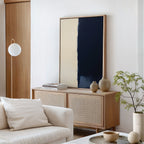 A minimalist living room with a light wood sideboard, neutral decor, a round white lamp, the Navy Division Canvas Art - Vertical in beige and dark blue, a white sofa with a cushion, and a small potted plant exudes modern elegance.
