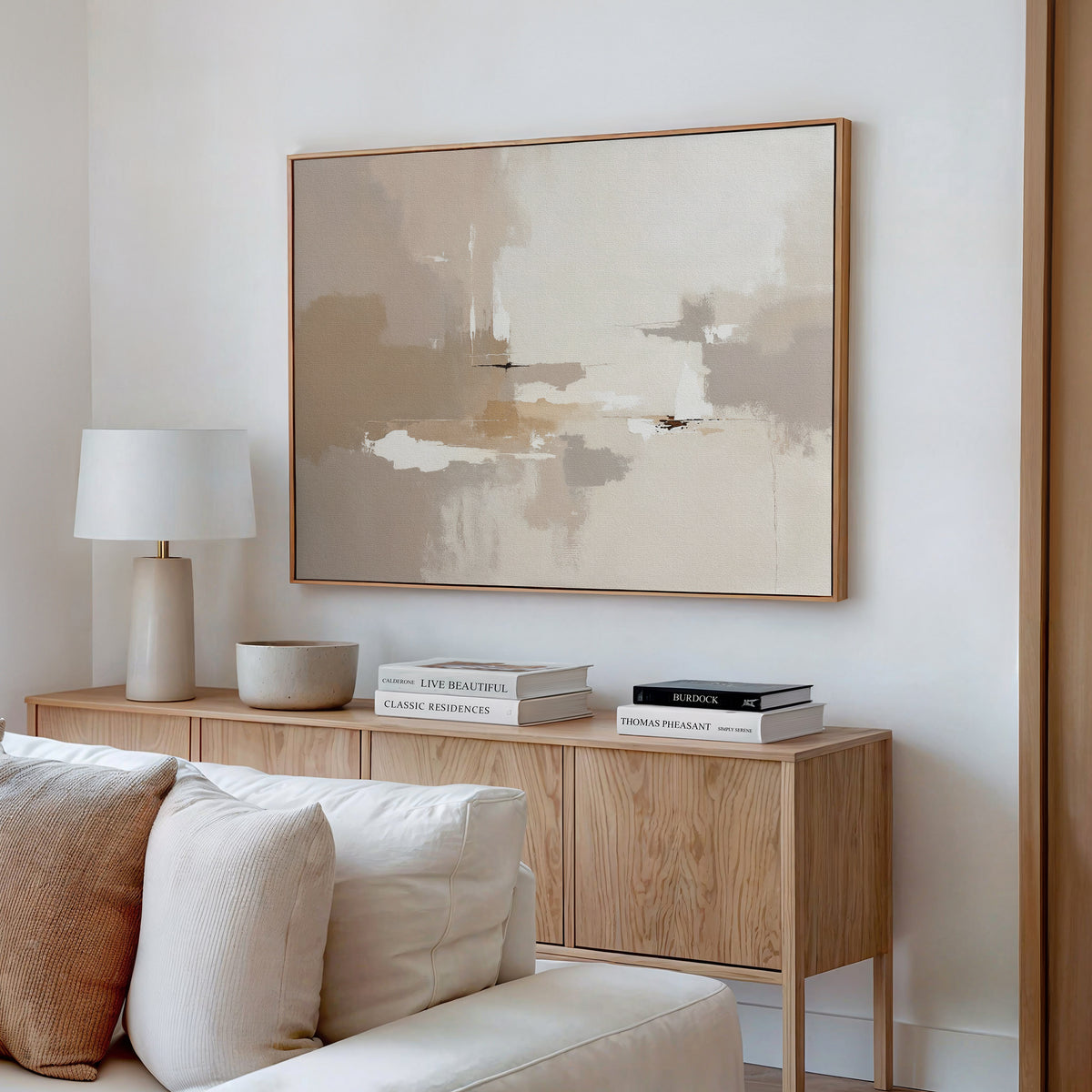 A modern living space showcases the Neutral Territory Canvas Art - Horizontal above a cream sofa with neutral cushions, complemented by a light wood sideboard, stacked books, a white lamp, and a bowl for contemporary elegance.