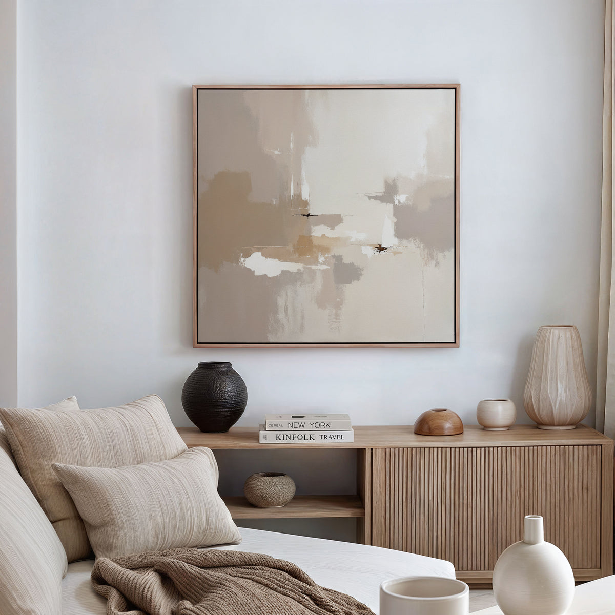 A minimal, modern living room features a beige sofa, neutral pillows, a wooden sideboard, vases, stacked books, and the Neutral Territory Canvas Art - Square in soft earth tones on a white wall. Natural light from the right enhances its contemporary elegance.