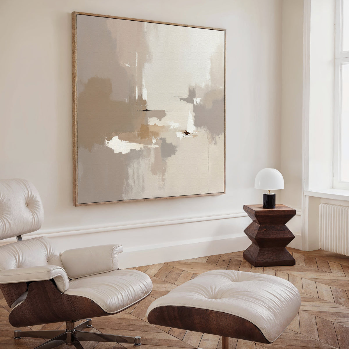 A modern living room with a cream lounge chair and ottoman, a Neutral Territory Canvas Art - Square in neutral tones, a geometric wooden side table, and a small white table lamp by a large window.