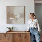 A woman in a white shirt and jeans leans against a wooden cabinet, admiring the Neutral Territory Canvas Art - Square on the wall, while a small plant and books add modern, minimalist style to the contemporary room.