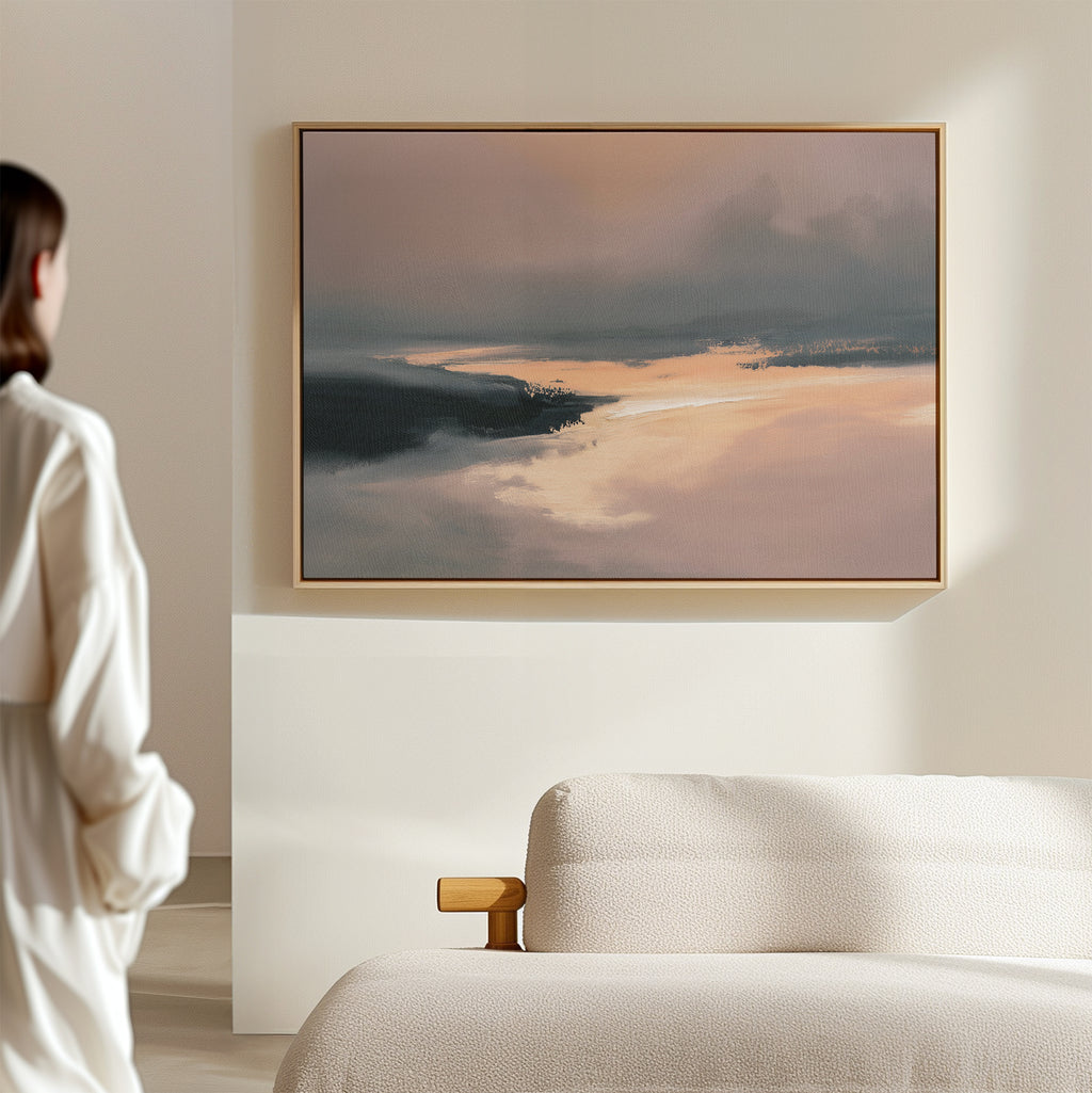 A person in a white robe admires the Nordic Fjord Canvas Art - Horizontal, featuring soft pink, orange, and blue hues, displayed above a textured white sofa in a minimal, light-filled room.