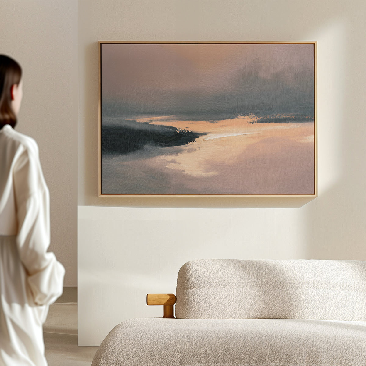 A person in a white robe admires the Nordic Fjord Canvas Art - Horizontal, featuring soft pink, orange, and blue hues, displayed above a textured white sofa in a minimal, light-filled room.