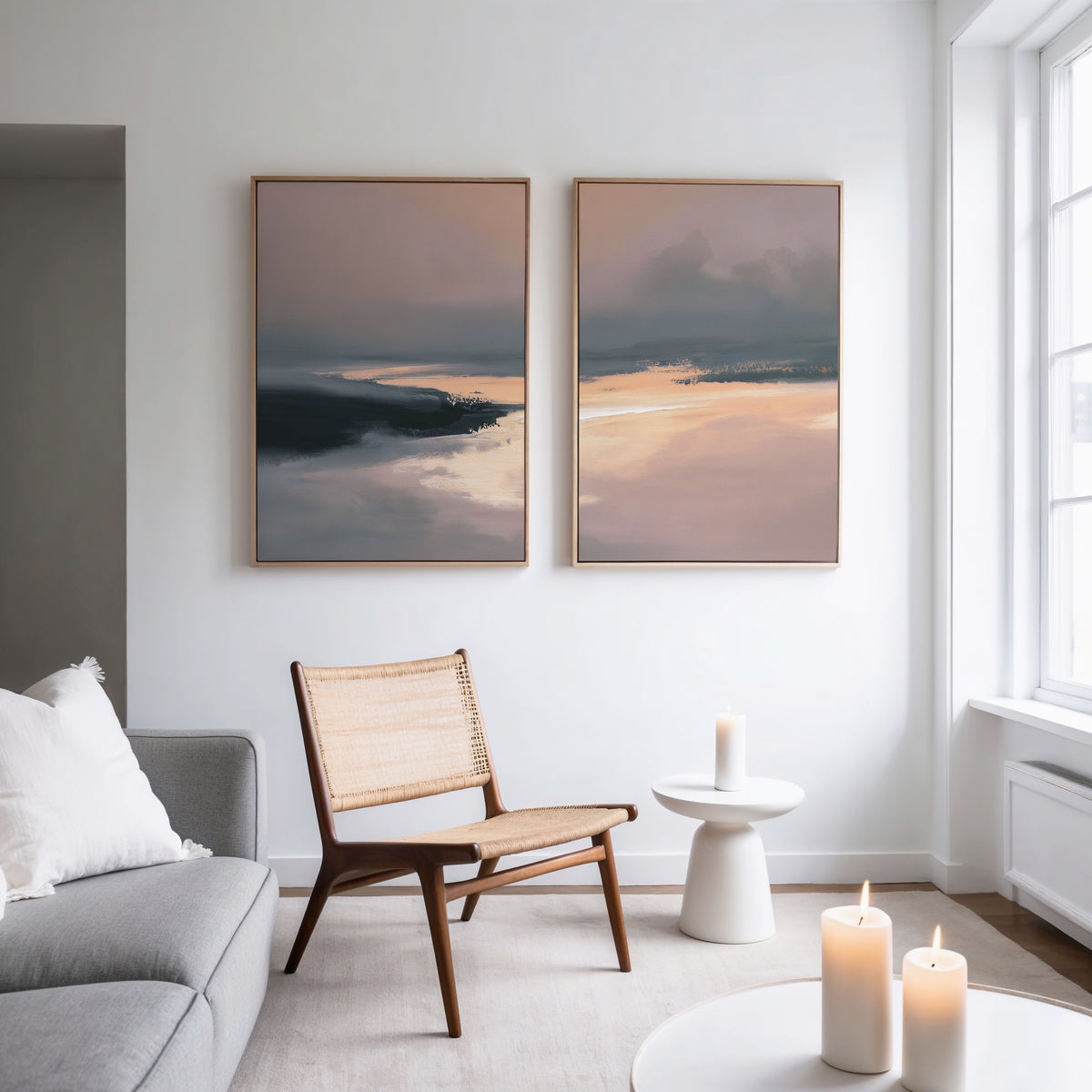 A modern living room with a wooden chair, small round table, lit candles, light gray sofa, and the Nordic Fjord - Set Of 2 abstract landscape wall art on a white wall by a large window.
