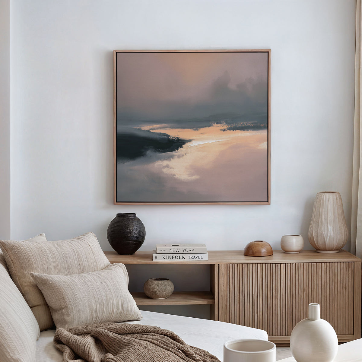 A modern, minimalist living room features a beige sofa, neutral cushions, wood sideboard with stacked books and decorative vases, and the Nordic Fjord Canvas Art - Square in soft earth tones as the focal wall décor.