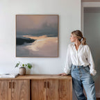 A woman in a white blouse and jeans stands by a wooden sideboard, admiring the Nordic Fjord Canvas Art - Square with blue, gray, and golden tones. A potted plant and books nearby complete this serene display.