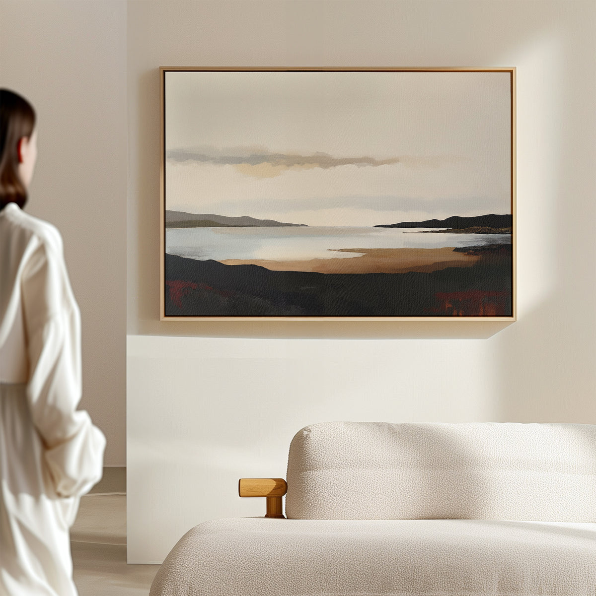 A person in a white shirt stands by a beige sofa in a minimalist room, viewing the Nordic Waters Canvas Art - Horizontal—a serene landscape of a lake and hills—displayed on a light-colored wall.