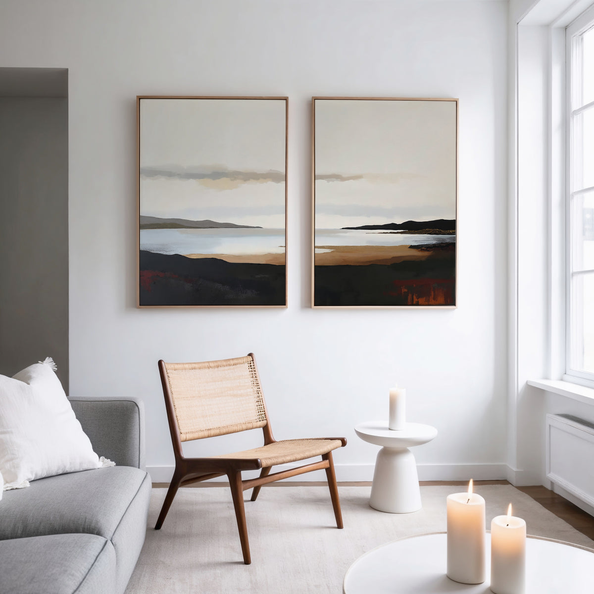 A modern, minimalist living room features a wooden chair, a side table with a candle, a light gray sofa, and calming decor. The Nordic Waters - Set Of 2 prints hang above as three lit candles glow near a large window.