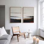 A modern, minimalist living room features a wooden chair, a side table with a candle, a light gray sofa, and calming decor. The Nordic Waters - Set Of 2 prints hang above as three lit candles glow near a large window.